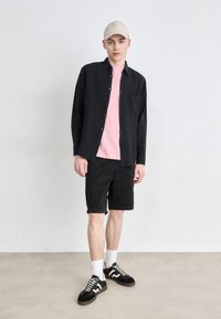 Camicia nera a bottoni sopra una maglietta rosa, pantaloni corti neri, calzini bianchi, sneakers nere e beige, e un cappellino beige chiaro.