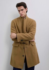 Hombre vestido con un abrigo largo color camel con botones negros, de pie con los brazos cruzados frente a un fondo liso de color gris claro.