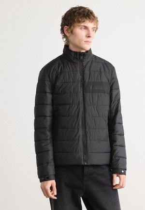 Black padded jacket with high collar, quilted texture, and zip fastening. Features a small logo patch on the chest and buttoned cuffs.