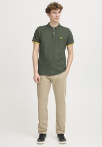 Grøn polo med gule accenter på ærmerne og et lille logo. Bæret med lysebeige bukser og sorte sneakers.