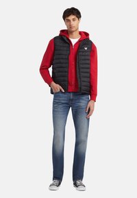 Chaleco acolchado negro sobre una sudadera con capucha roja, combinado con vaqueros azules y zapatillas negras. El chaleco tiene cierre de cremallera y textura acolchada.