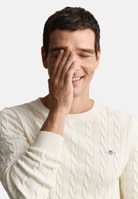 Maglione crema a trecce con polsini a coste; mano che copre parzialmente il viso, mostrando un leggero sorriso; texture morbida e design classico.