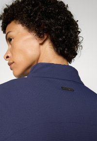 Navy blue textured shirt with a subtle diamond pattern. Features a stand collar and a black metal brand tag at the back.