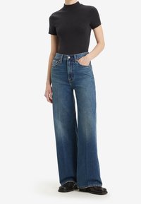 Femme portant un col roulé noir à manches courtes, un jean bleu taille haute à jambes larges, des chaussures noires, debout avec une main dans la poche.