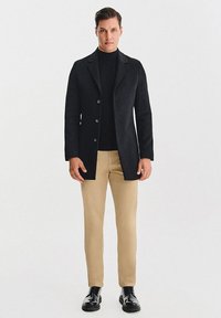 Czarny wełniany płaszcz z wyciętymi klapami, jednorzędowy z pięcioma guzikami, w zestawieniu z czarnym golfem i beżowymi spodniami. Błyszczące czarne buty.