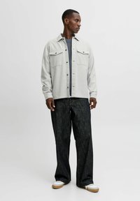 Camisa de botones gris claro con dos bolsillos en el pecho, llevada sobre una camiseta de color azul marino, combinada con jeans oscuros de pernera ancha y zapatillas blancas.