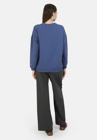 Sudadera azul con cuello redondo y mangas largas, combinada con pantalones de pierna ancha en gris oscuro y zapatos rojos, vista desde la espalda.