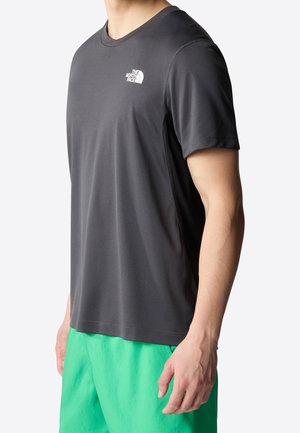 Mand iført en mørkegrå kortærmet trænings-T-shirt med hvidt North Face-logo og lysegrønne shorts, stående mod en ensfarvet baggrund.