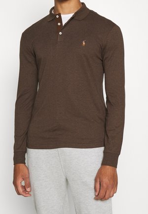 Homme portant un polo marron à manches longues avec un petit logo brodé et un pantalon de survêtement gris clair, debout les bras relâchés.