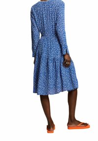 Femme portant une robe midi bleue à motifs à manches longues et des tongs orange, debout, dos tourné vers l’avant.