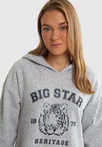 Šviesiai pilkas per didelis džemperis su priekiniu tigro grafiniu vaizdu ir navy blue spalvos tekstu "BIG STAR 1979 HERITAGE". Minkšta medžiaga, su raišteliu reguliuojamu gobtuvu.