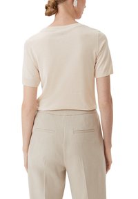 Jasno beżowa koszulka z krótkim rękawem o okrągłym dekolcie, wykonana z miękkiego materiału. W zestawie z jasnobeżowymi, eleganckimi spodniami z tylnymi kieszeniami.