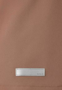 Bronzefarbenes Gewebe mit glatter Textur, das ein gesticktes Etikett mit silbernem Rand und der Prägung "name it" in der Mitte aufweist.