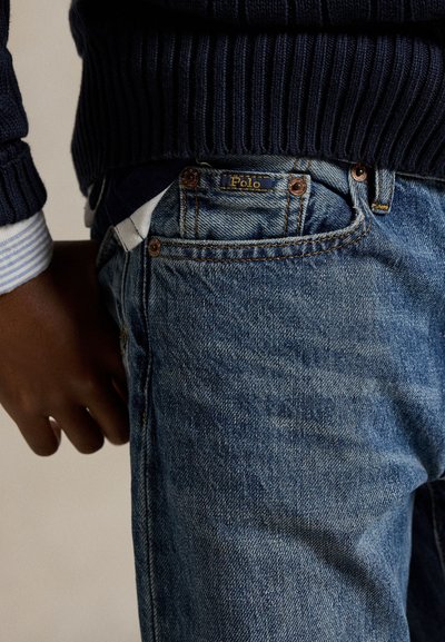 Közeli kép egy személyről, aki kék farmert visel Polo címkével és egy sötétkék pulóvert egy csíkos ing mandzsettája fölött.