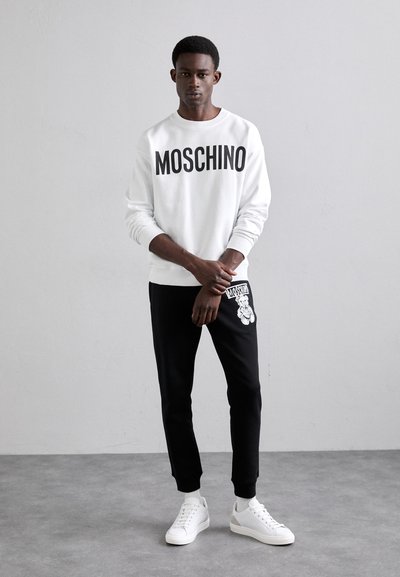 Sudadera blanca con "MOSCHINO" en texto negro en negrita, combinada con pantalones jogger negros que presentan un gráfico de un oso, complementada con zapatillas blancas.