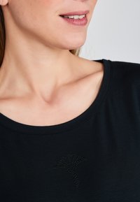 Haut noir à manches courtes avec un décolleté large, présentant un design de logo texturé en strass sur le devant. Texture de tissu lisse.