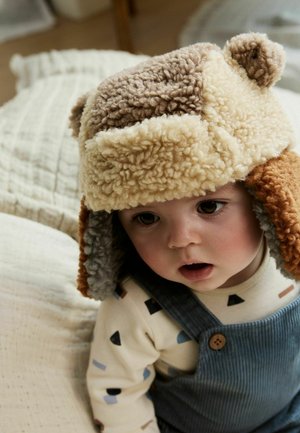 Toddler wearing a fluffy, multi-colored winter hat with ear flaps and a cream shirt with patterned shapes, sitting on a textured white blanket.