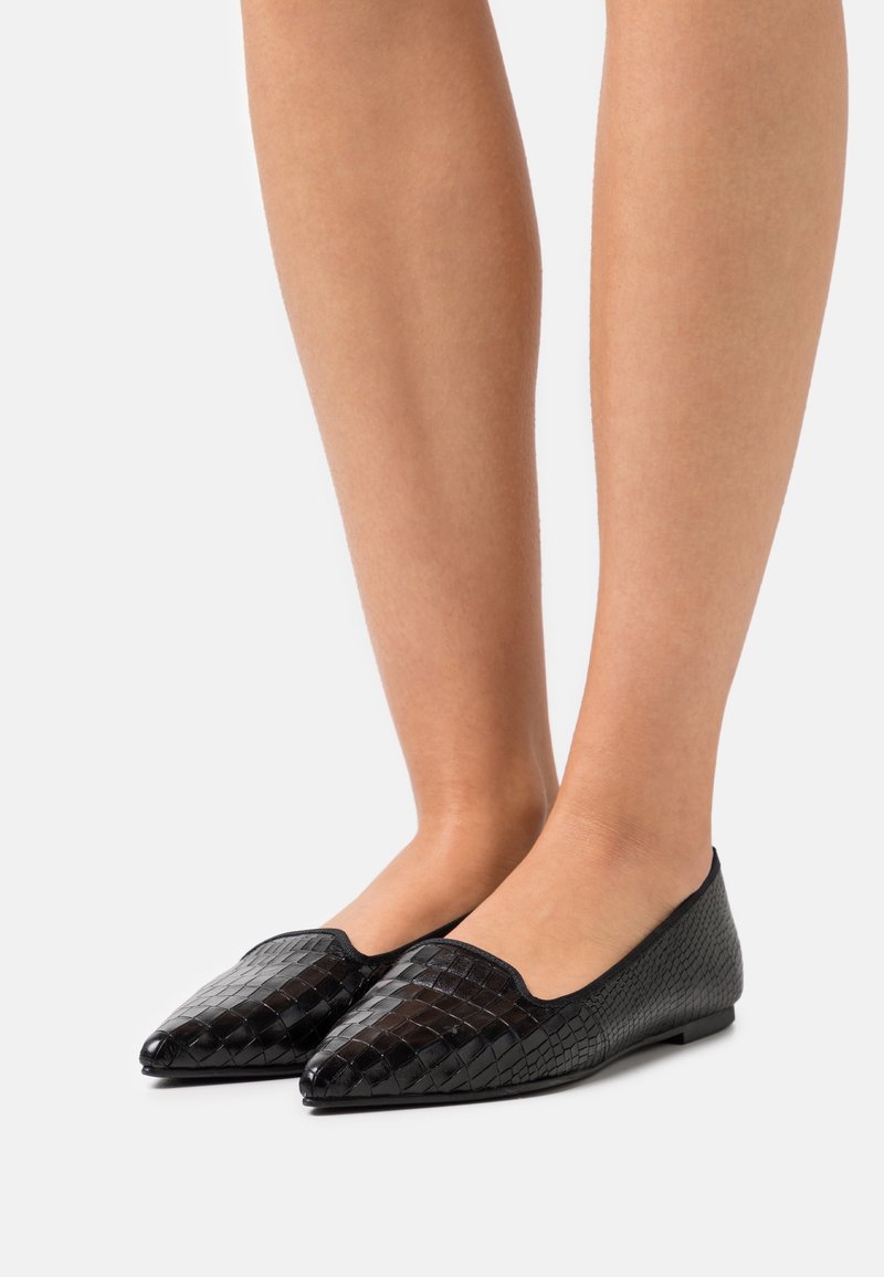Jambes portant des ballerines noires à bout pointu avec un motif tissé texturé sur un fond uni clair.
