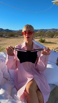 Musta brasiliinalainen rintaliivi ohuilla olkaimilla käsissä; malli pukeutunut pitkään, raidalliseen vaaleanpunaiseen napitettavaan paitaan. Aurinkoinen aavikkotausta.