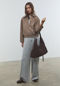 Giacca marrone con zip e colletto, pantaloni sportivi grigi a gamba larga e una borsa a tracolla in suede marrone scuro. Scarpe marroni completano il look.
