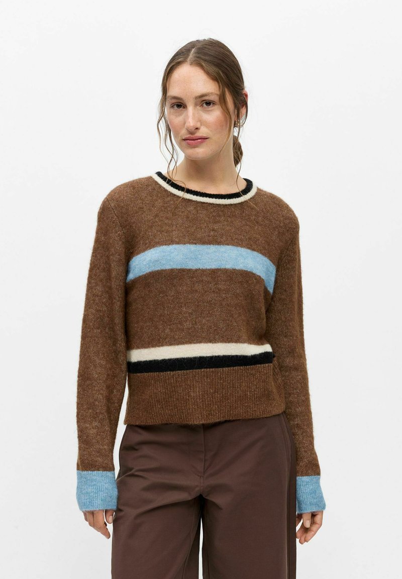 Jeune femme aux cheveux bruns porte un pull marron avec des rayures bleues, blanches et noires ainsi qu'un pantalon marron sur un fond uni.