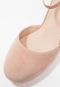Chaussure plate en daim rose clair avec un bout arrondi et un design découpé. Dotée d'une fine bride à la cheville et d'une doublure intérieure lisse.