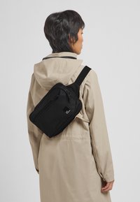 Schwarze Umhängetasche aus strapazierfähigem Stoff, mit rechteckiger Form, zwei Reißverschlüssen und einem Logo auf der Vorderseite, diagonal über einem beigen Mantel getragen.