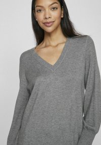Maglione grigio a V realizzato in un tessuto morbido; maniche lunghe con vestibilità ampia e polsini a costine, caratterizzato da una texture liscia e da un design minimale.