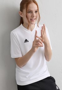 Vörös hajú lány fehér Adidas pólóban és fekete nadrágban, az ujjait számolja, egy fehér textúrájú falnak támaszkodva, mosolyog.