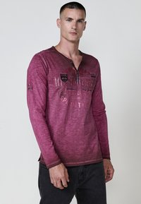 Burgundi színű, hosszú ujjú ing henley nyakkivágással. Visszafogott textúrájú anyag, nyomtatott grafikák és oldalsó hasíték jellemzi. Sötét farmernadrággal viselve.
