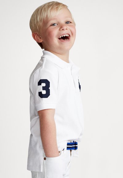 Biele polokošeľa s tmavomodrým číslom 3 na rukáve, spojená s bielymi nohavicami a viacfarebným pásom s pruhmi. Dieťa sa usmieva a pozerá doprava.