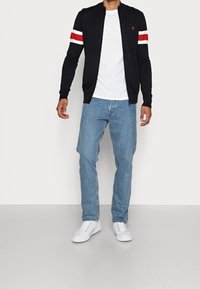 Svart zip-up hoodtröja med röda och vita randiga detaljer på ärmarna, vit t-shirt, ljusblå jeans och vita sneakers.