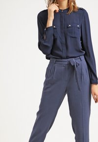 Blusa blu navy con maniche lunghe, polsini con bottoni, due tasche sul petto, abbinata a pantaloni larghi dello stesso colore e una cintura annodata in vita.