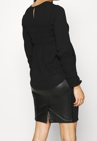 Černý top s dlouhým rukávem a zvrásněnou texturou na zadní straně, doplněný černou koženou sukní s rozparkem vzadu.
