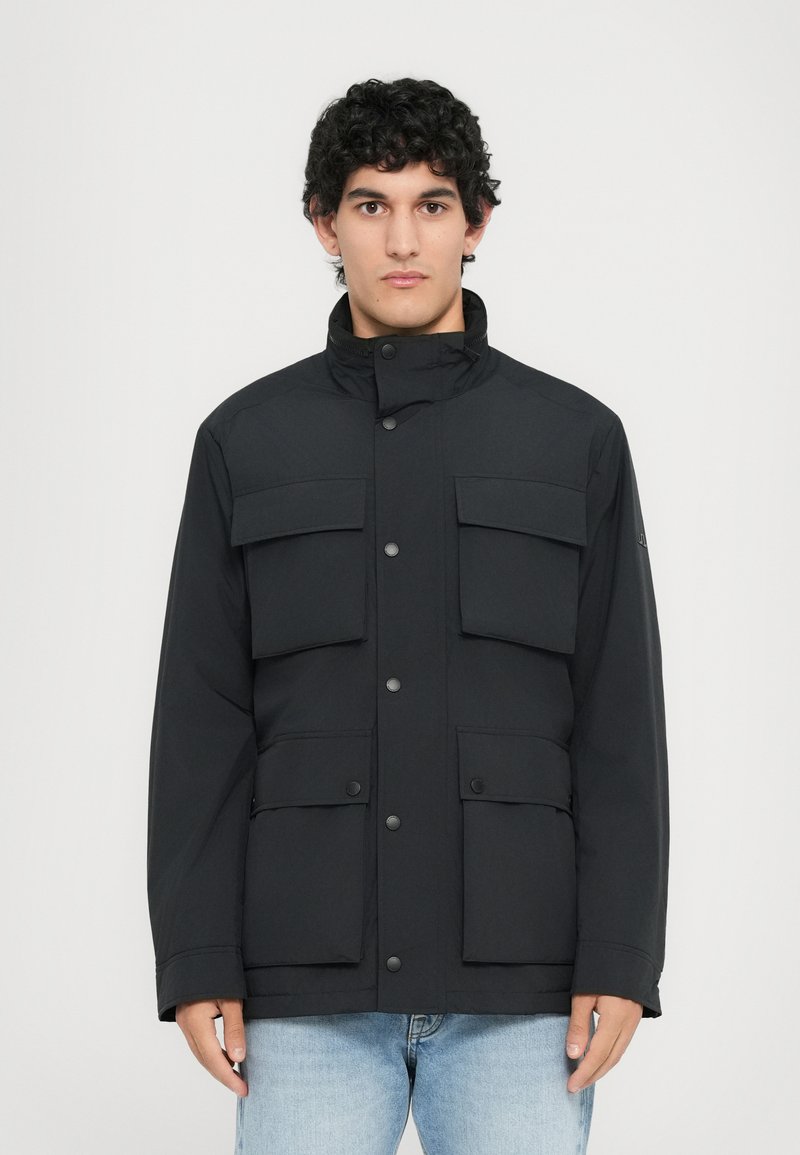 Uomo con capelli ricci e scuri che indossa una giacca nera con quattro tasche frontali e jeans azzurri chiari, in piedi davanti a uno sfondo bianco semplice.