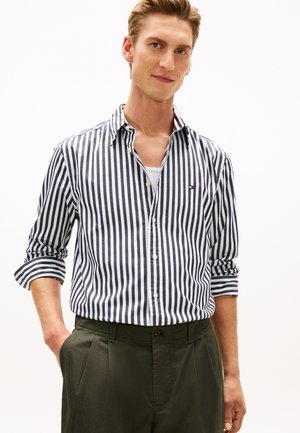 Muž nosící černo-bílou vertikálně pruhovanou košili s knoflíky, s vyhrnutými rukávy, zastrčenou do tmavě zelených kalhot, stojící s jednou rukou v kapse.