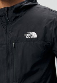 Sort zipjakke lavet af et glat, syntetisk materiale. Har en hætte og et hvidt logo på venstre bryst. Inkluderer en lynlåslomme.