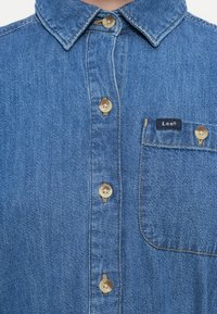 Nærbilde av en blå denimskjorte med beige knapper, spiss krage og brystlomme som har et lite Lee-merkelapp.