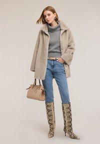 Beige faux bontjas over een grijs gebreide trui, gecombineerd met een blauwe spijkerbroek. Accessoires zijn onder andere een beige handtas en kniehoge boots met slangenprint.