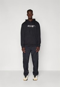 Sweatshirt preta com capuz, bolso frontal e detalhe gráfico. Combinada com calças escuras, largas e ténis bege com detalhes pretos.