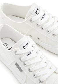 White canvas sneakers with a textured finish, featuring silver eyelets, flat laces, and a logo label on the tongue.