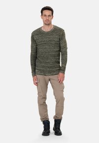 Camisa de manga larga verde jaspeada con cuello redondo, combinada con pantalones cargo beige y botas negras con cordones, de pie sobre un fondo blanco.