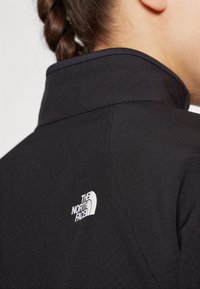 Crni jakna od tkanine s visokim ovratnikom, s bijelim logotipom "The North Face" na lijevom ramenu i suptilnom teksturiranom površinom.