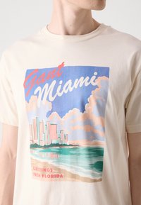 Beež puuvillane T-särk, millel on värviline Miami graafika, sealhulgas hooned, pilved ja vesi. Tekst ütleb "Gant Miami" ja "Tere tulemast Floridasse."
