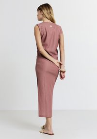 Woman standing facing away in a sleeveless, ribbed, dusty pink midi dress with slide sandals and chunky bracelets on her wrist.