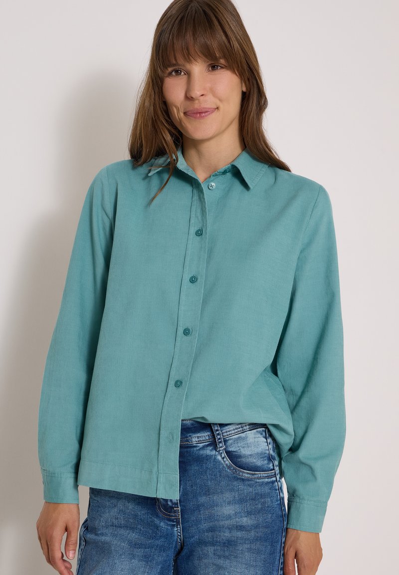 Femme aux cheveux bruns raides, portant une chemise bleu sarcelle à boutons et un jean bleu, souriant sur un fond clair.
