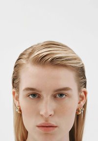 Pendientes de aro de oro con múltiples anillos entrelazados en un acabado liso y pulido, llevados por una modelo con cabello lacio y claro frente a un fondo neutro.