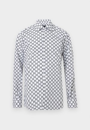 Bela srajca z dolgimi rokavi na gumbe z abstraktnim črnim geometrijskim vzorcem in klasičnim koničastim ovratnikom na enobarvnem ozadju.