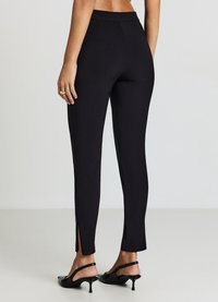 Schwarze, figurbetonte Hosen mit glatter Textur, die seitliche Schlitze am Saum und eine mittelhohe Taille aufweisen. Kombiniert mit schwarzen High Heels.