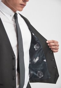 Mężczyzna w czarnym garniturze i krawacie, trzymający otwartą kurtkę, aby odsłonić wewnętrzne podszycie w kwiatowy wzór w odcieniach szarości i czerni.